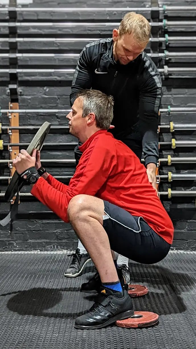 A client performing a heel-raised frog squat with a weight in their hands while Chris ensures they are in the correct position